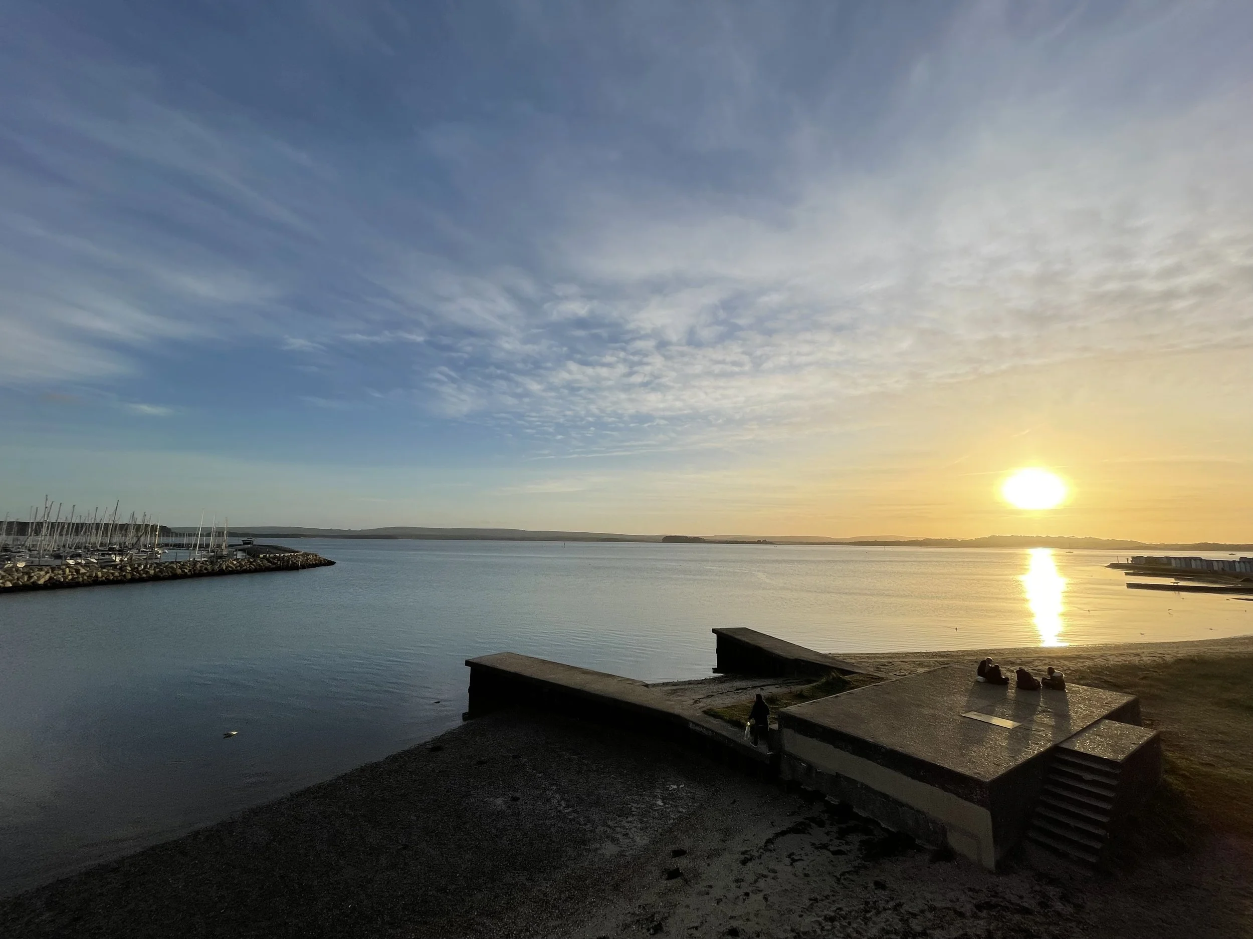 A sunset over a calm body of water with a partly cloudy sky, a marina on the left, a small dock, and people sitting on the shoreline.