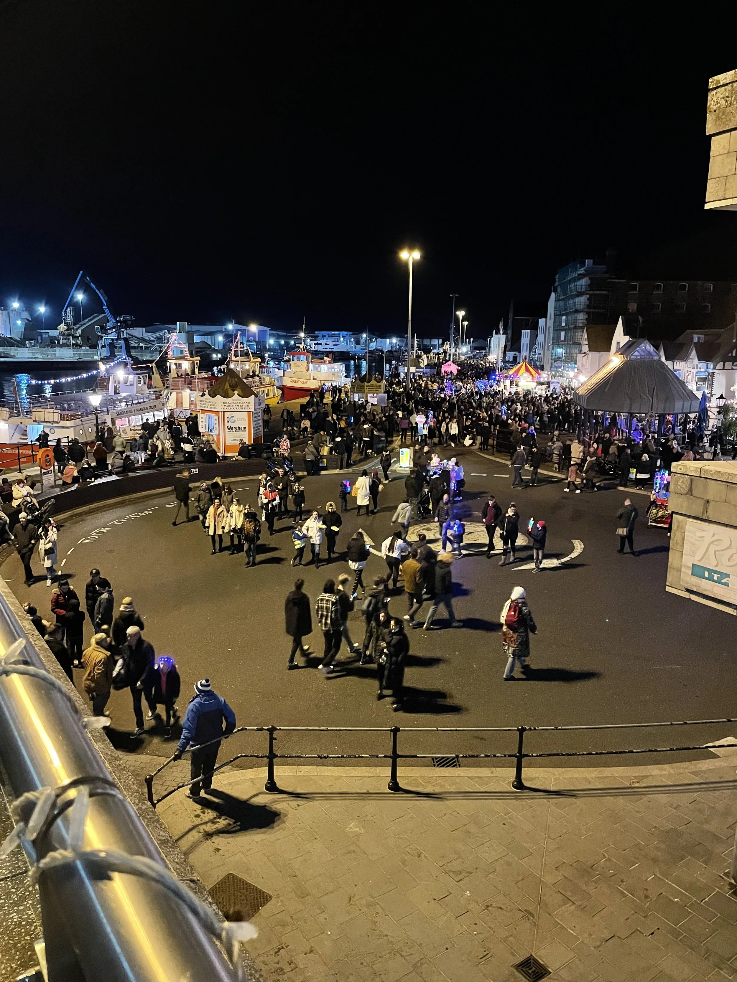 Night view of a crowded outdoor fair or festival with food stalls, a carousel, and people walking around.
