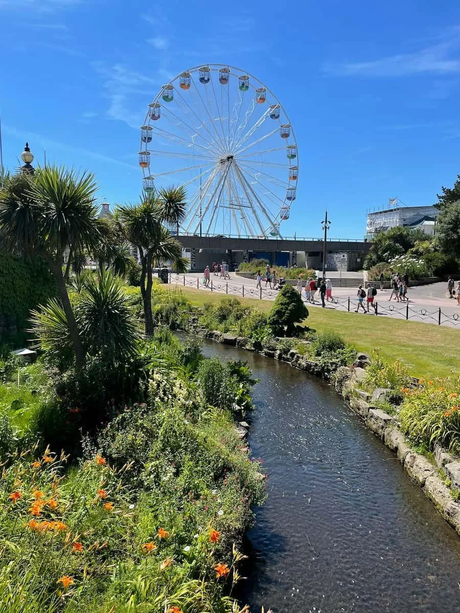 A large Ferris wheel in a park with people walking along a paved path. The scene includes lush greenery, a small stream, and bright blue sky.