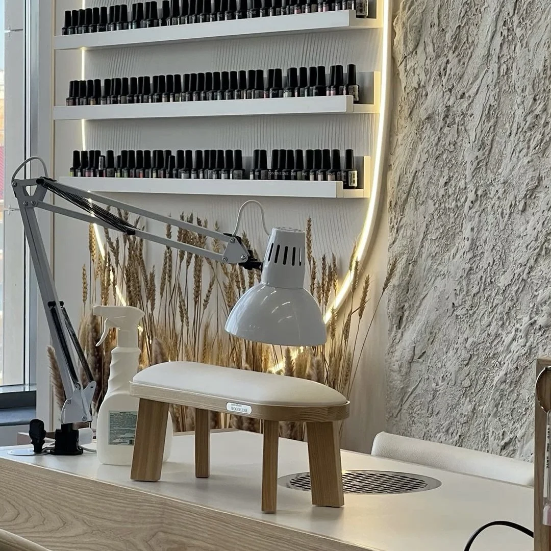 Interior of a nail salon with shelves of nail polish bottles on the wall, a white desk lamp, a spray bottle, and a small wooden stool on a light-colored desk, with decorative wheat stalks and textured wall in the background.