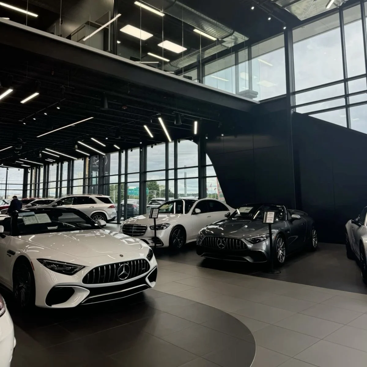 Interior of a car dealership showing various Mercedes-Benz cars on display inside a modern showroom with large glass windows and a black ceiling with embedded lighting.