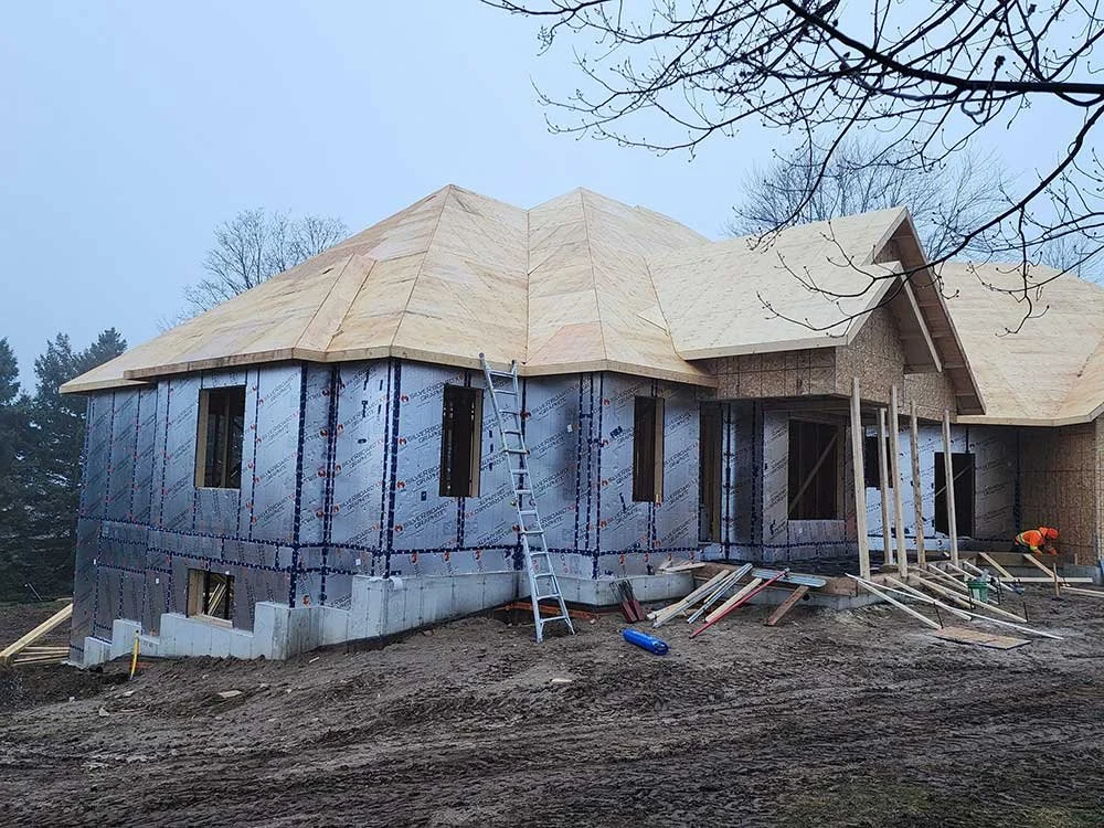 A house under construction with a partially completed wooden roof and insulated walls, surrounded by construction materials and tools on a dirt ground.