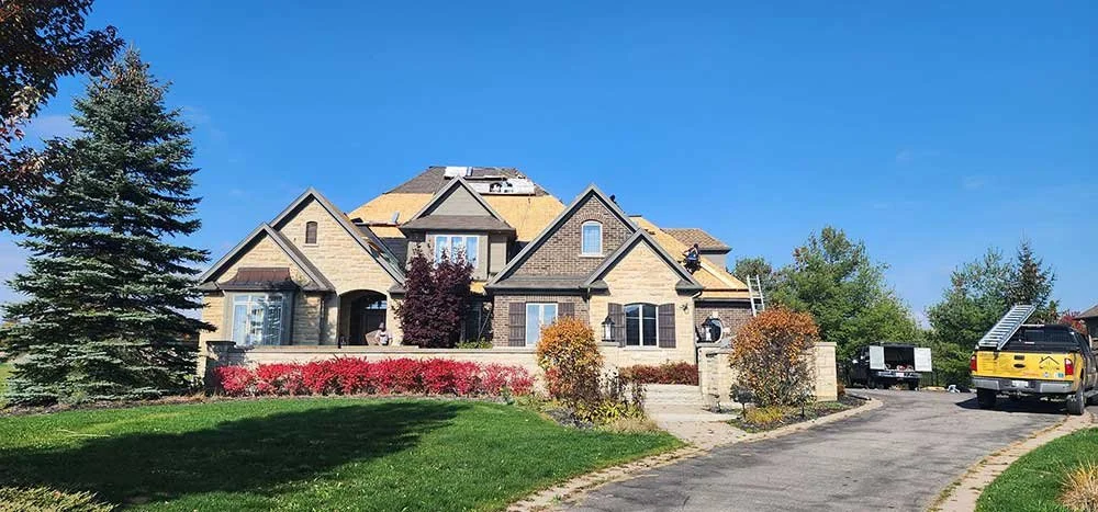 A house under construction with a partially completed roof, surrounded by a landscaped yard with trees and bushes, and a driveway with construction trucks parked.