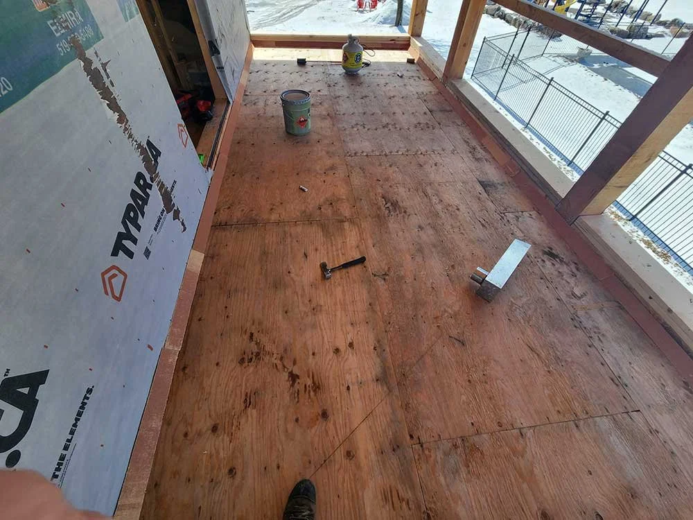 Construction site of a balcony or deck with plywood flooring, partially installed, with tools and materials like a hammer, wood block, paint can, and a spray bottle. There is a view of snow outside through safety railing.