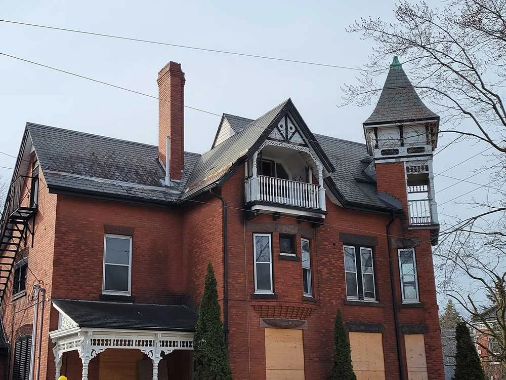 An old Victorian-style house with red brick walls, a turret with a pointed roof, and boarded-up windows and doors. Quality Care Roofing is a Kitchener-Waterloo roofing company and roof repair contractor.