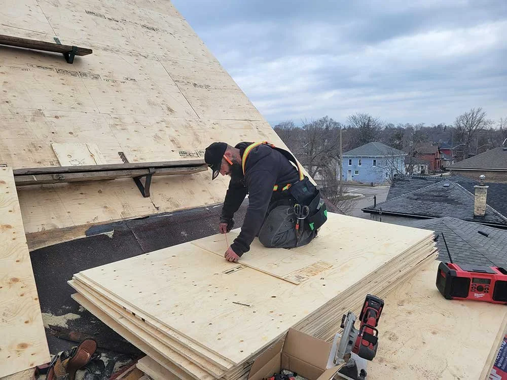A construction worker wearing safety gear, including a harness, kneeling on a pile of plywood sheets while working on a roof during daytime.