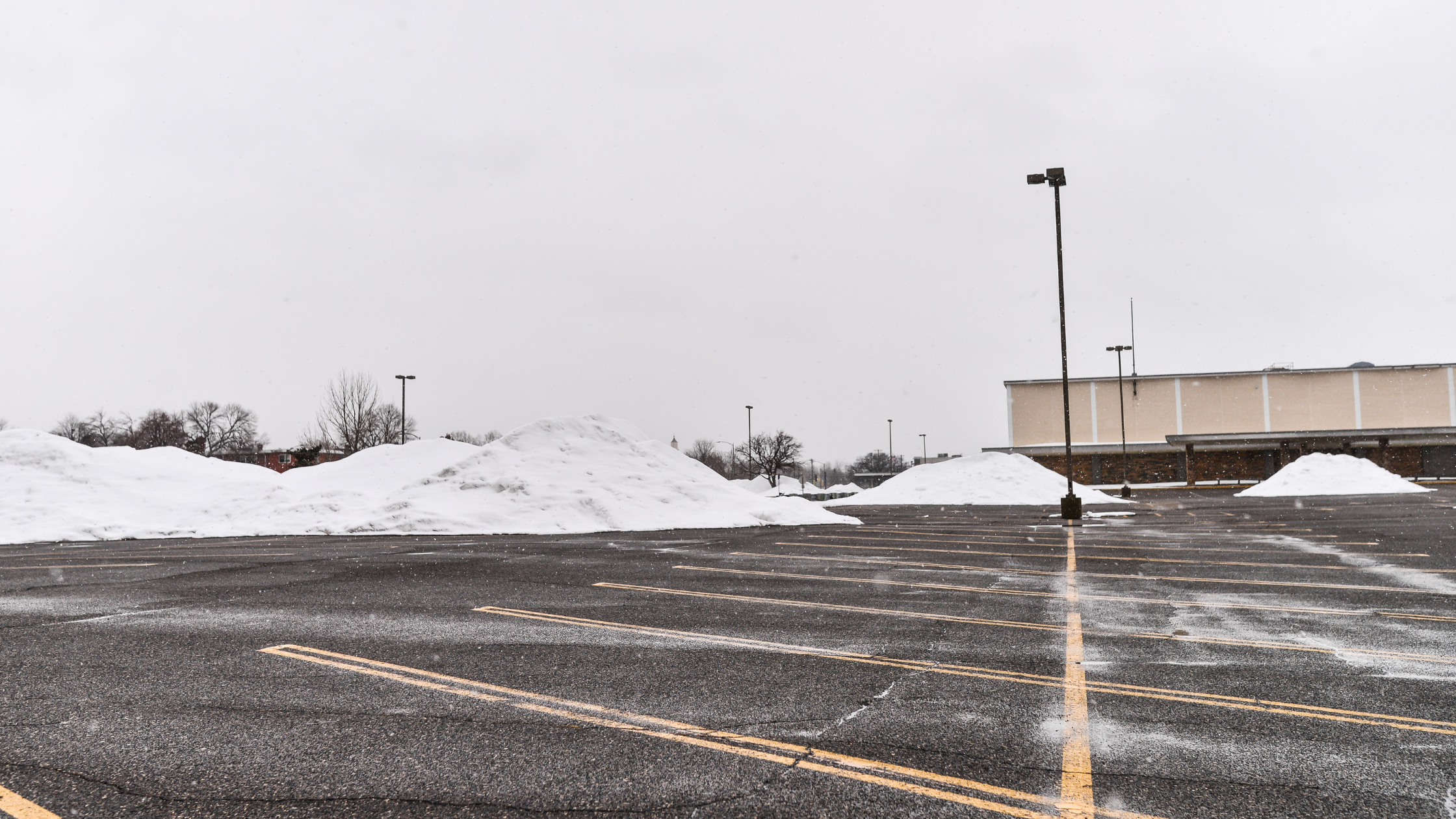 Empty parking lot with snow piles and a few light poles, overcast sky.