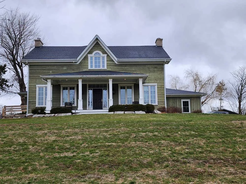 A large, two-story house with green siding, white trim, and black front door, featuring a front porch with white columns, situated on a grassy hill with a cloudy sky in the background.
