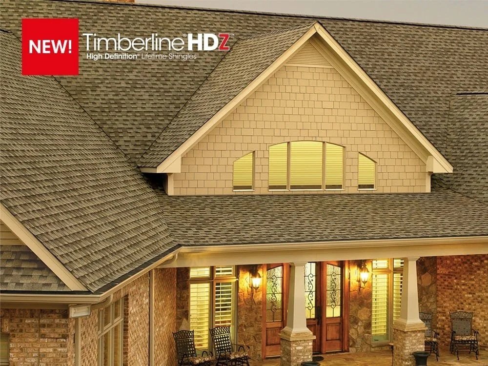 Close-up of a house's roof with gray shingles, brick walls, and a front porch with wooden front door and lantern-style lights.
