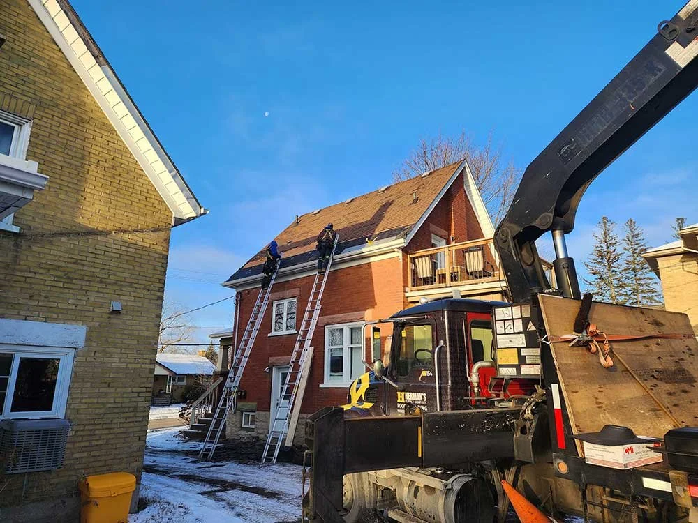Two workers on ladders cleaning or inspecting the roof of a red brick house, with a large truck equipped with a crane nearby. Snow is on the ground and the sky is clear.