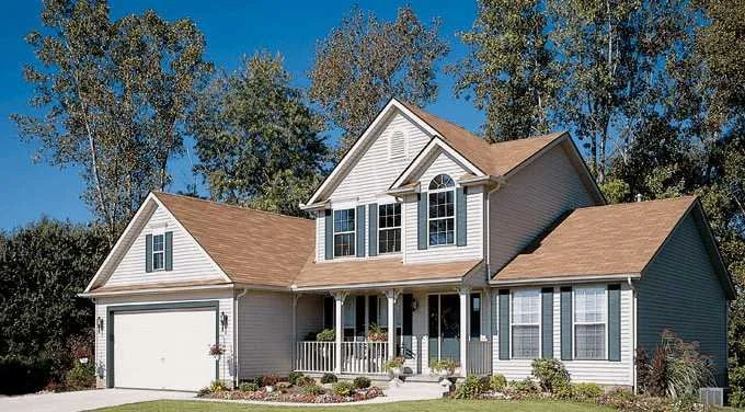 A two-story suburban house with beige siding, brown roof, white trim, front porch, and attached garage, surrounded by trees and a lawn.