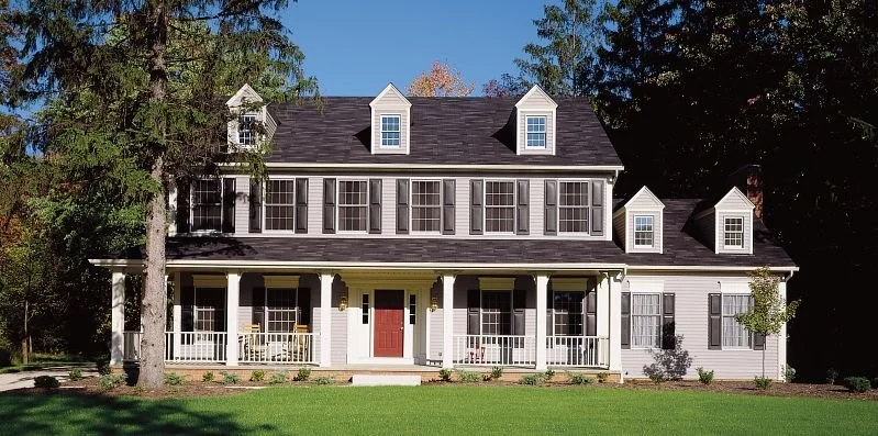 Large two-story house with white siding, black shutters, a covered front porch, and dormer windows on the roof, surrounded by trees and a green lawn.