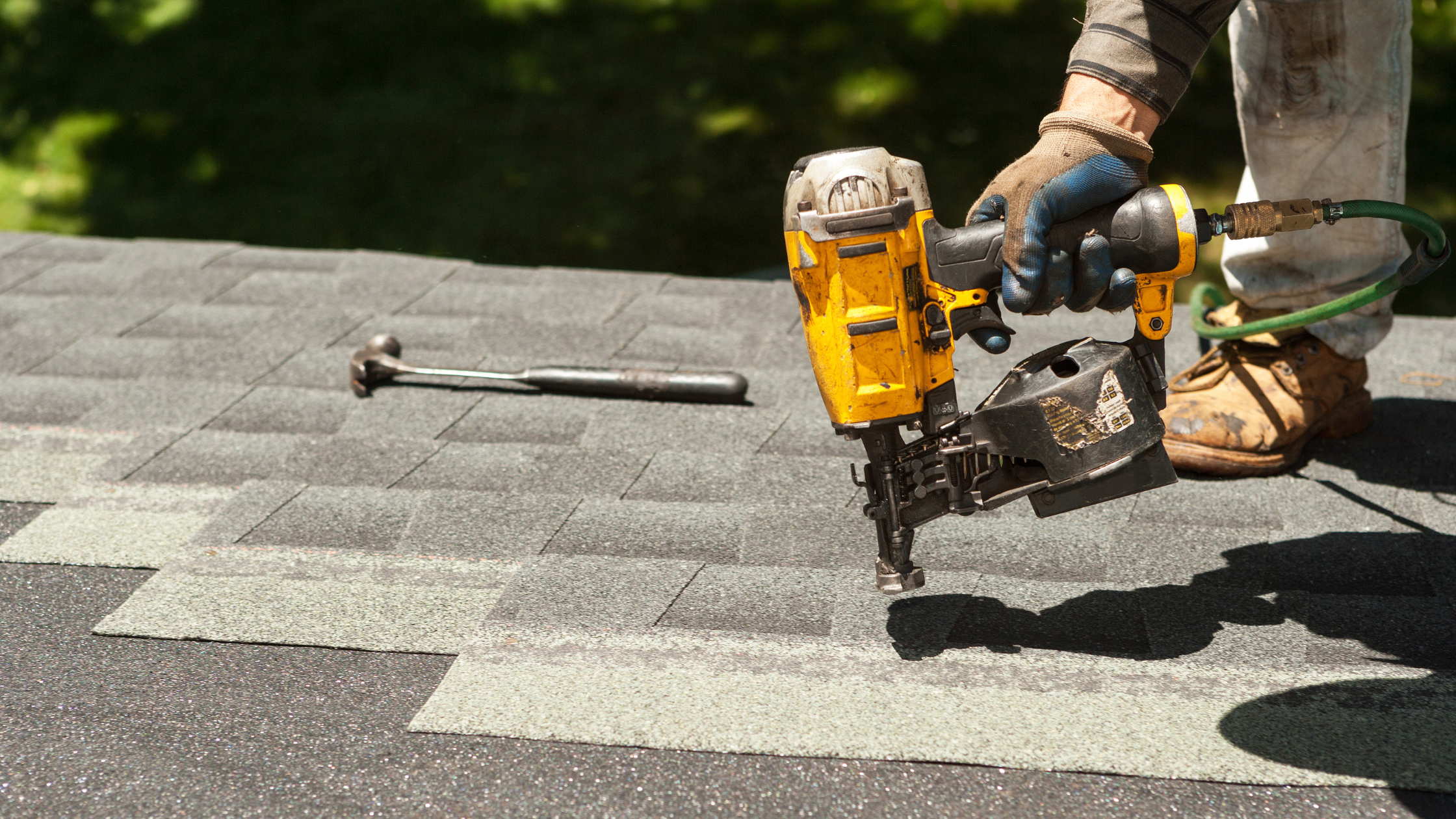 A worker installing asphalt shingles on a roof using a pneumatic nailer. The worker is wearing gloves, work boots, and is kneeling on the roof.