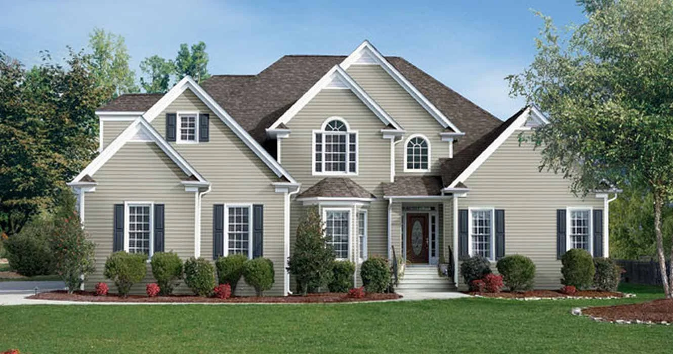 Large suburban house with beige siding, multiple gabled roofs, white trim, and dark shutters, surrounded by well-maintained lawn and bushes.