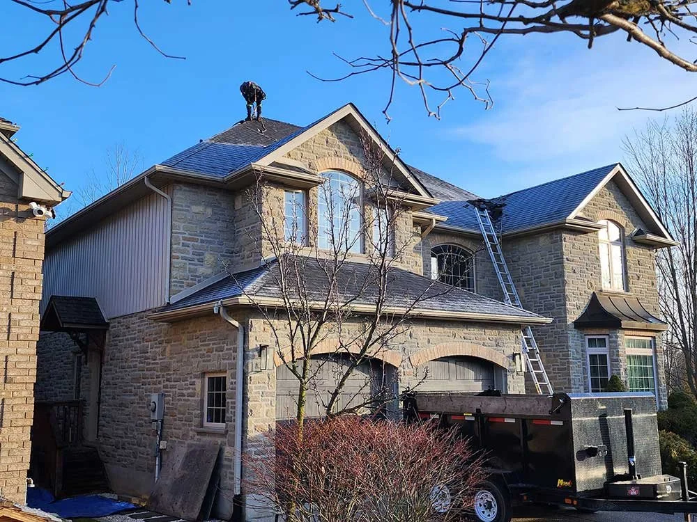 A two-story house with stone and siding exterior, blue roof, and a ladder leaning against the upper floor, with a worker on the roof.