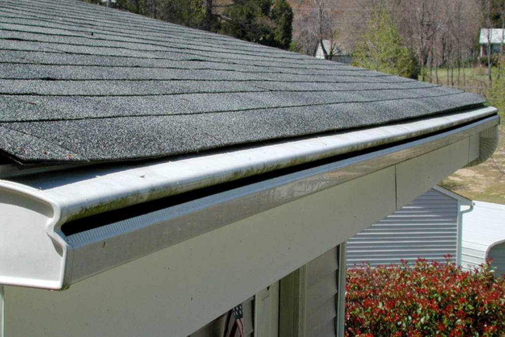 Close-up view of a house roof with asphalt shingles, gutter system, and some trees and houses in the background.