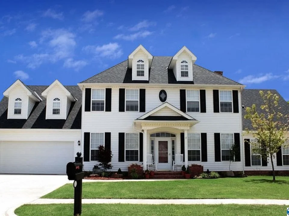 A large, white, two-story house with black shutters and a gabled roof, front porch with steps, surrounded by a well-maintained lawn and trees on a sunny day.