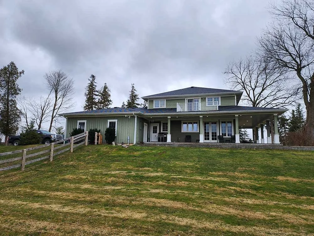 A large two-story house with green siding, a covered porch, and outdoor seating, situated on a grassy hill with a fence and trees in the background under gray cloudy skies.