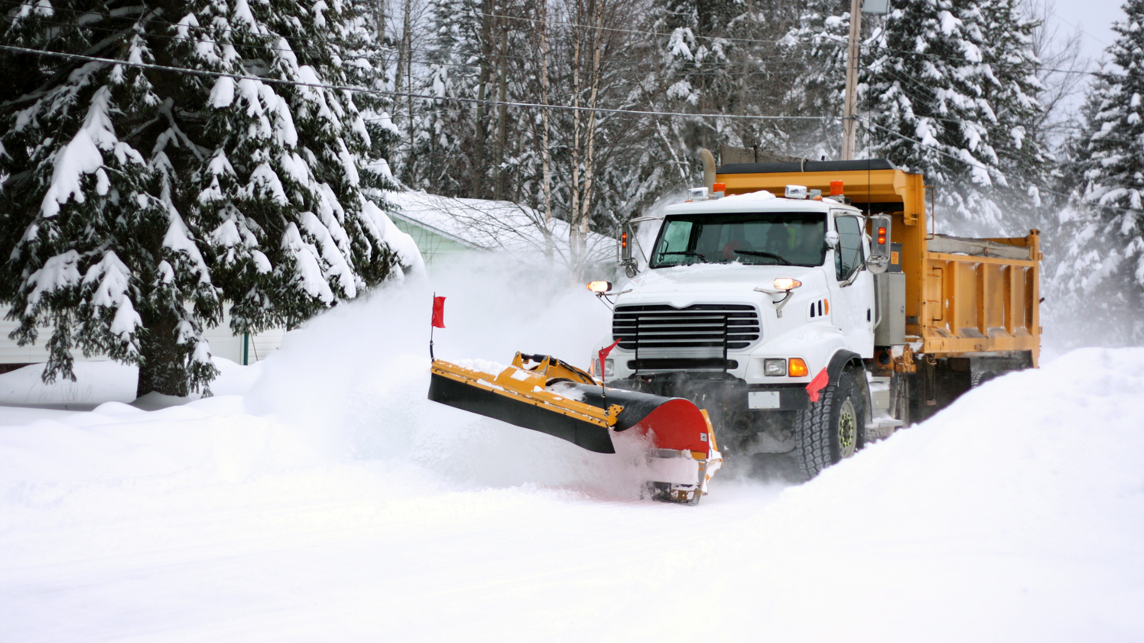A front view of a yellow snow plow clearing snow from a road in a snowy landscape with trees