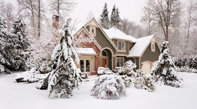 A snowy suburban house with a gray exterior, brick accents, and a chimney, surrounded by snow-covered trees and bushes.