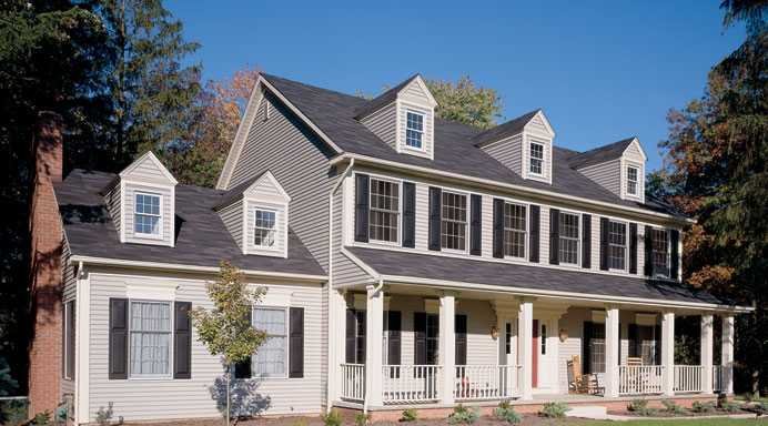 Large two-story house with gray siding, black shutters, multiple dormer windows, a front porch, and surrounded by trees