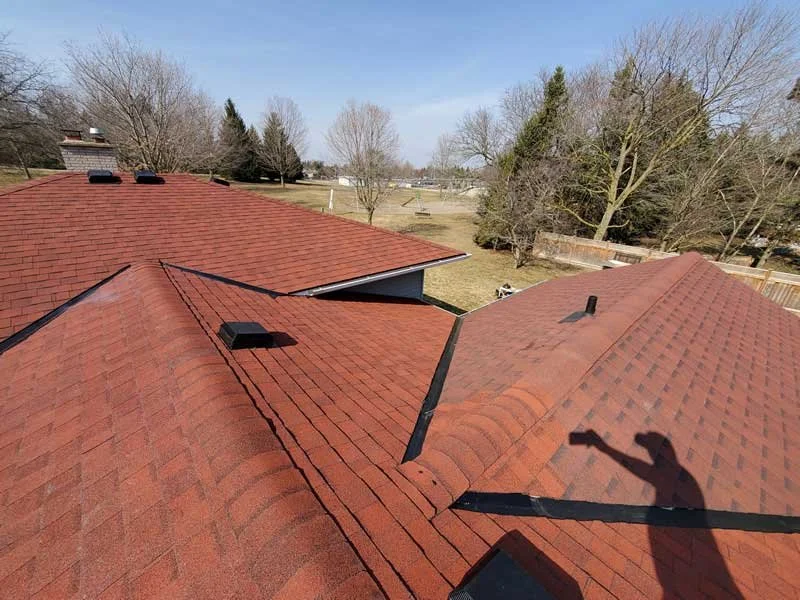 View of a house roof with red shingles and multiple vents, with a backyard and trees in the background.