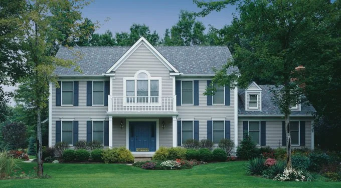 Large two-story house with white siding, blue shutters, and a front porch, surrounded by green lawn and trees.