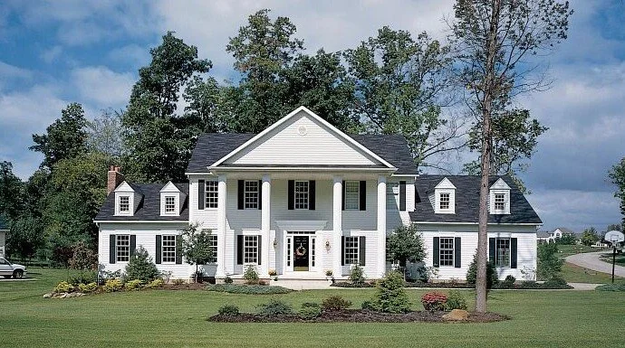 Large white two-story house with classical columns at the front entrance, surrounded by a landscaped yard with trees and bushes, under a partly cloudy sky.