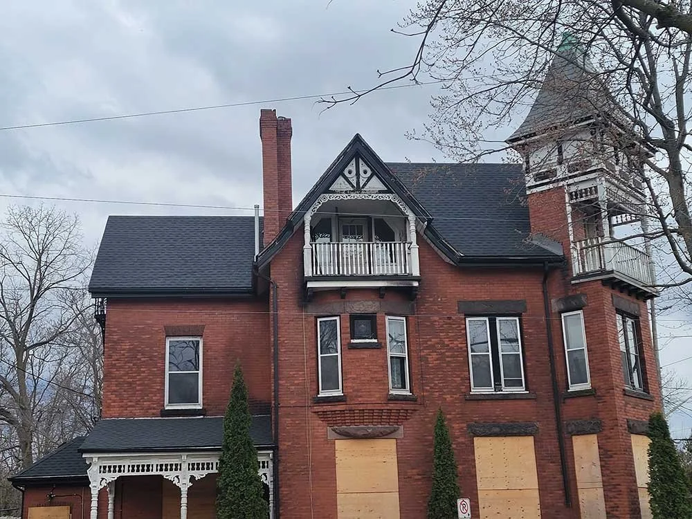 An old, large, brick house with boarded-up windows and decorative woodwork on the front porch. The house has multiple stories, including a tower-like structure on one side, and appears to be vacant or under renovation. The sky is overcast and there a