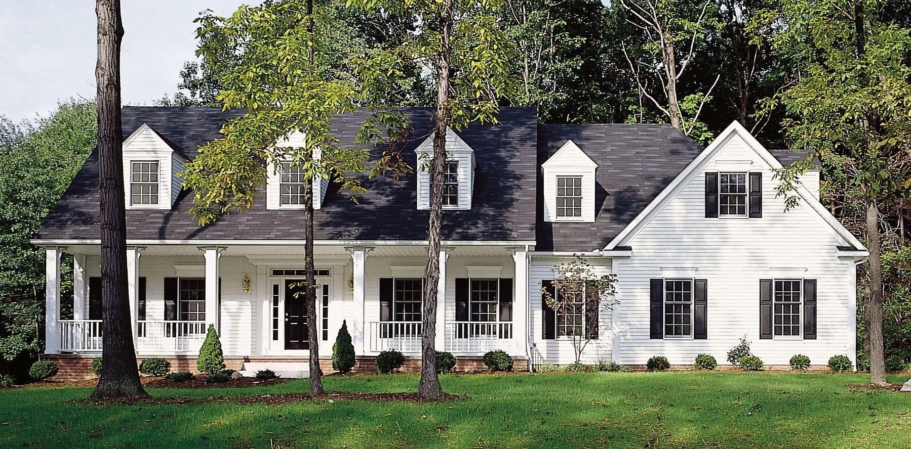 White two-story house with black shutters, front porch, dormer windows, surrounded by a green lawn and trees.
