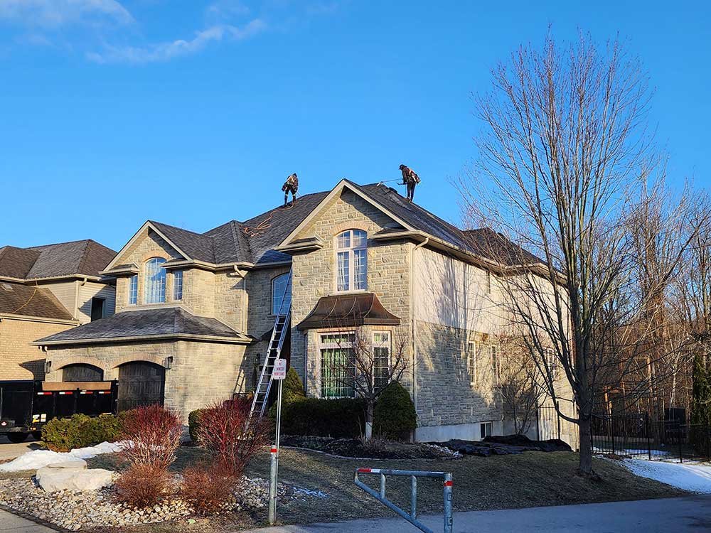 Two construction workers on the roof of a large stone house working on roof repairs or installation, with a ladder leaning against the front of the house, and leafless trees in the yard. Quality Care Roofing in Kitchener-Waterloo, Ontario, Canada.