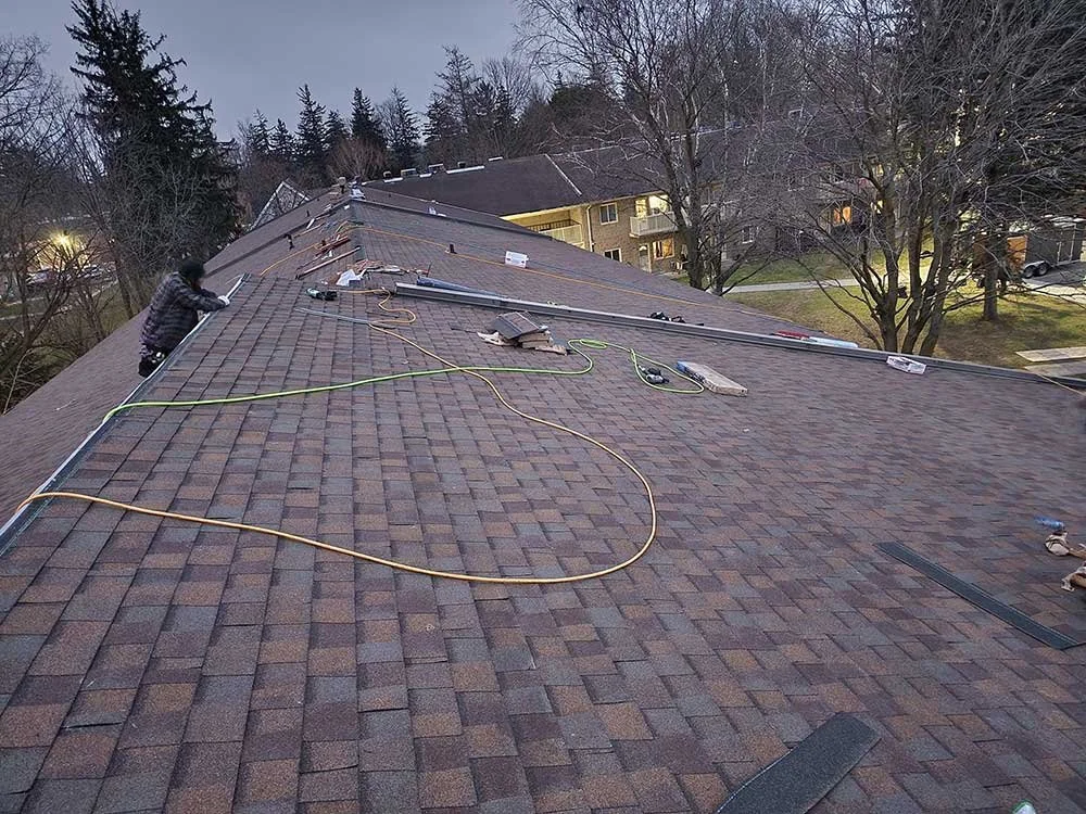 A worker installing solar panels on a sloped roof of a residential building during dusk, with trees and neighboring houses visible in the background.