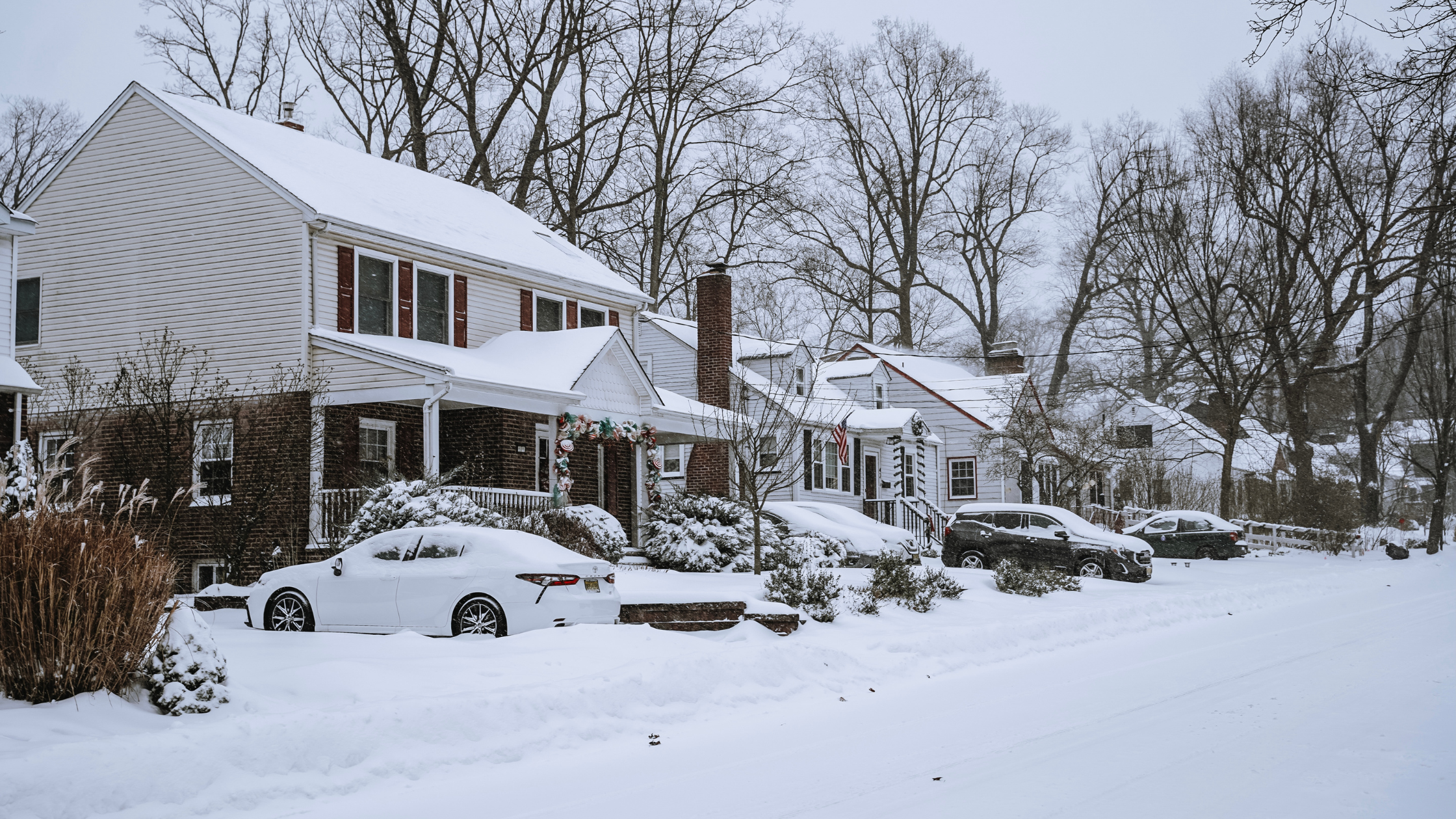 Snow-covered suburban street with houses, cars, and leafless trees in winter.