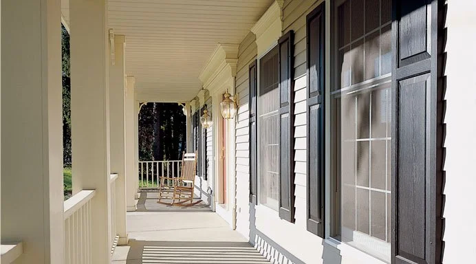 Front porch of a house with black shutters, rocking chair, and a railing, illuminated by sunlight.