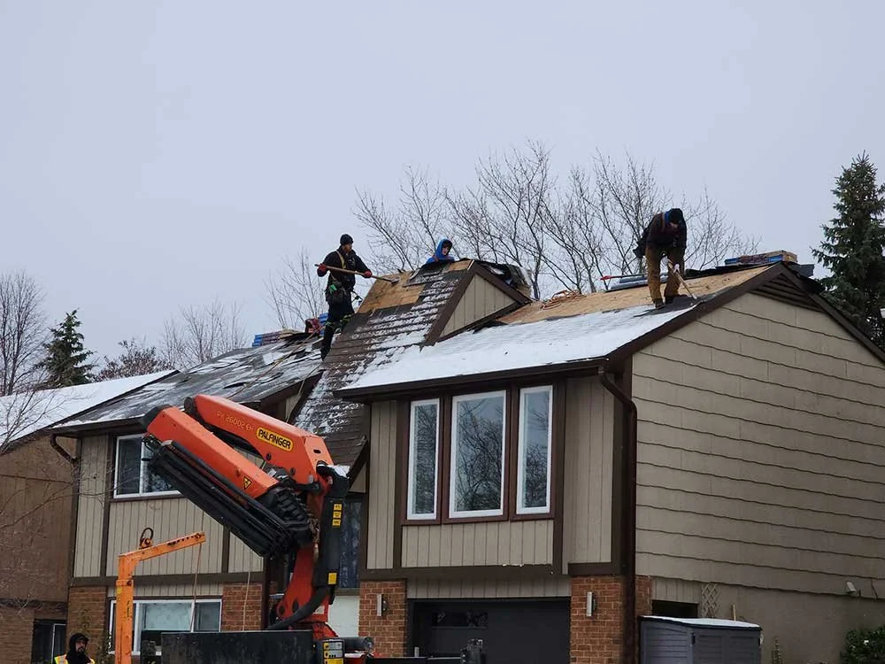 People working on a house roof with snow, using a crane to lift roofing materials or tools.