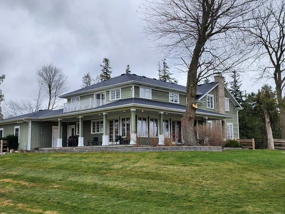 Large two-story house with light green exterior, white trim, and a front porch with chairs. There is a large tree in front, and the yard is grassy with a slight incline. Trees without leaves are visible in the background.