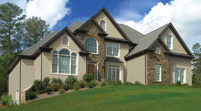Large two-story house with beige siding, stone accents, multiple gabled roofs, and large windows, situated on a grassy hill under a partly cloudy sky.