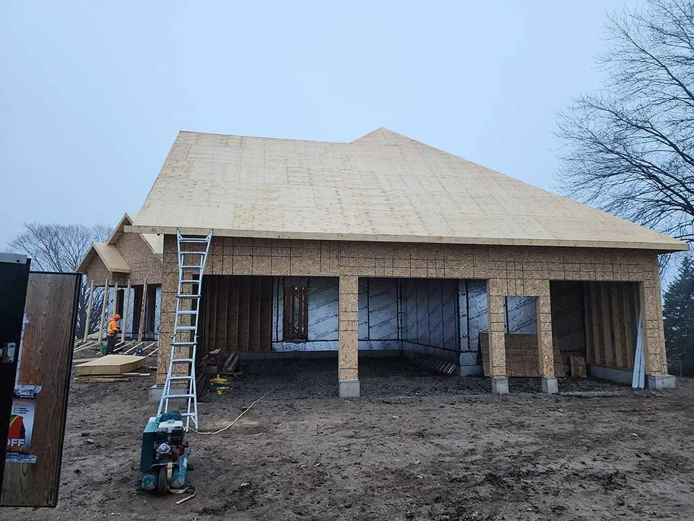 Under construction house with exposed wooden framing, ladder, construction worker, and construction equipment on dirt ground.