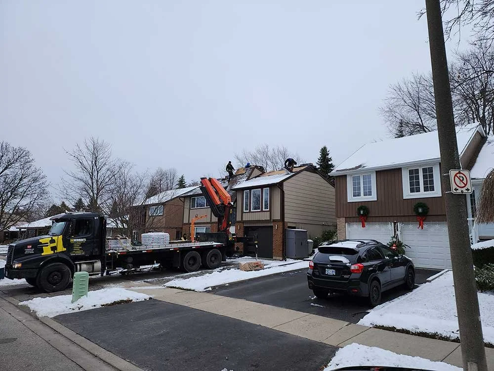 Construction workers on rooftops of suburban houses installing or repairing the roofs, with a flatbed truck and crane nearby during winter, with snow on the ground and Christmas wreaths on the garage doors.