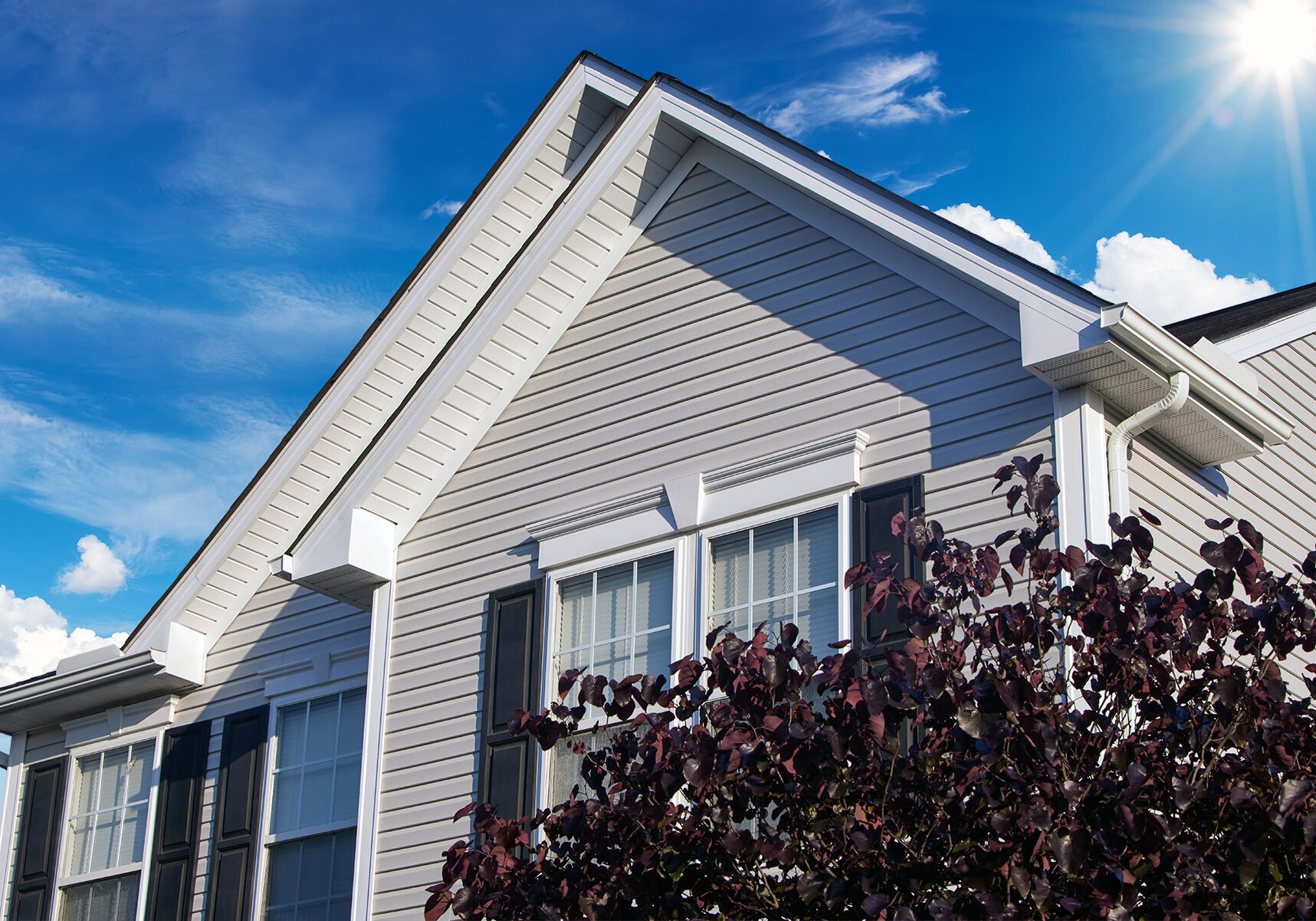 Close-up of a white house with vinyl siding, black window shutters, and a gabled roof. The sky is blue with some clouds, and the sun is shining. Dark purple-leaved shrubbery is in front of the house.
