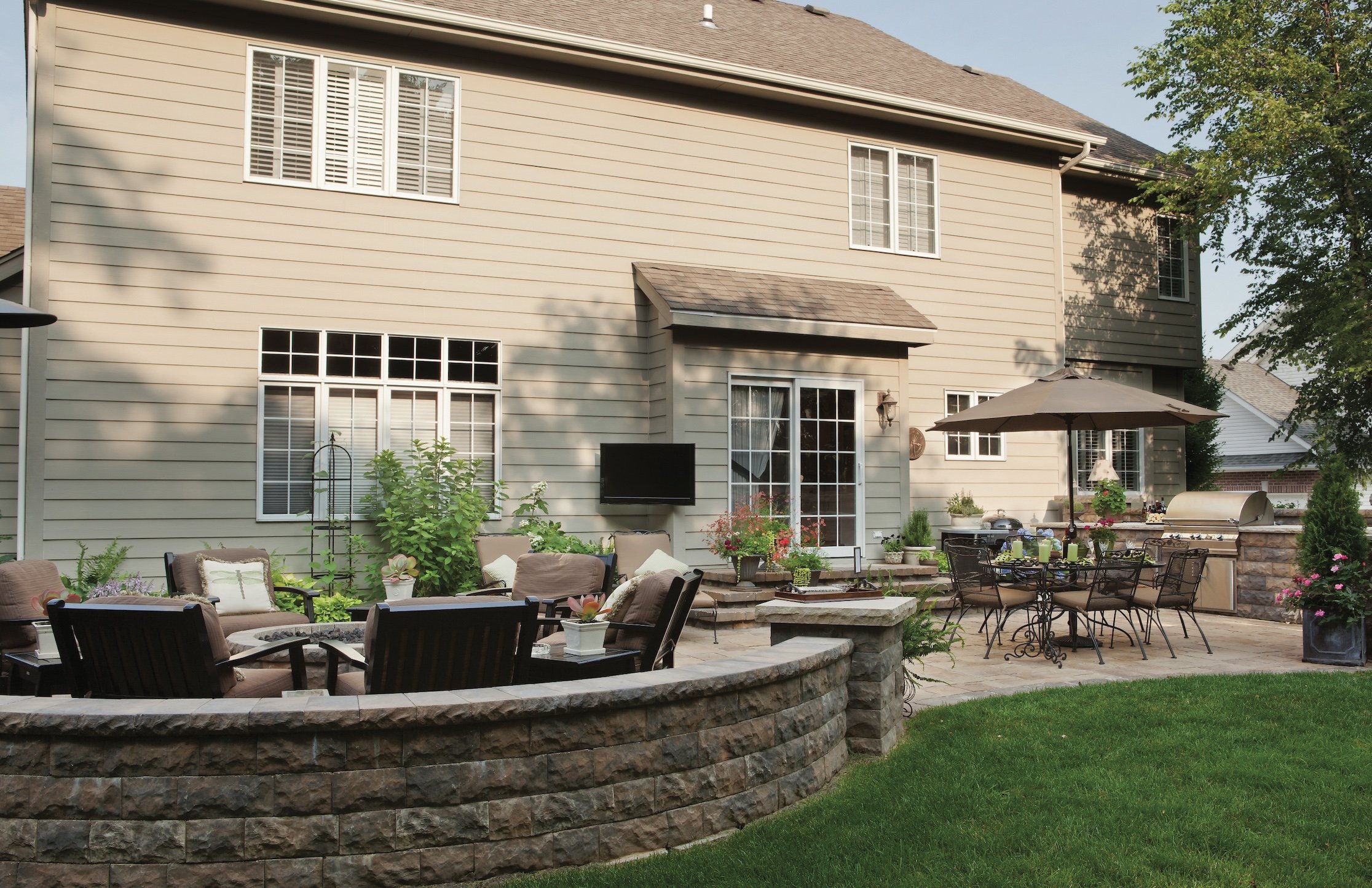 Backyard patio with seating area, table under umbrella, outdoor grill, potted plants, and a stone retaining wall in front of a beige house with large windows.