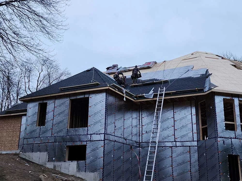 Workers installing roofing on a house under construction with a ladder leaning against the structure.