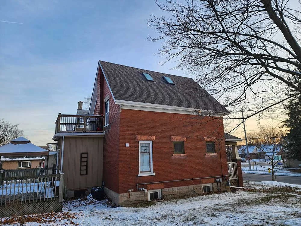 A brick house with a brown shingled roof, multiple skylights, and a small backyard deck with a ladder, in winter with some snow on the ground and leafless trees.