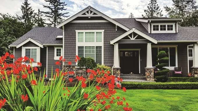 Front view of a gray house with a gable roof, white trim, and a small porch, surrounded by a well-maintained lawn and red flowers in the foreground.