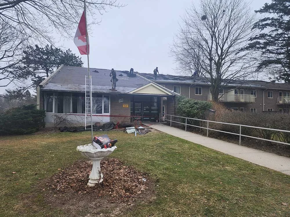 Residential building with roof damage from roofers working on the roof. A flagpole with a Canadian flag, a birdbath, and a wheelchair ramp are in the front yard. Overcast sky.
