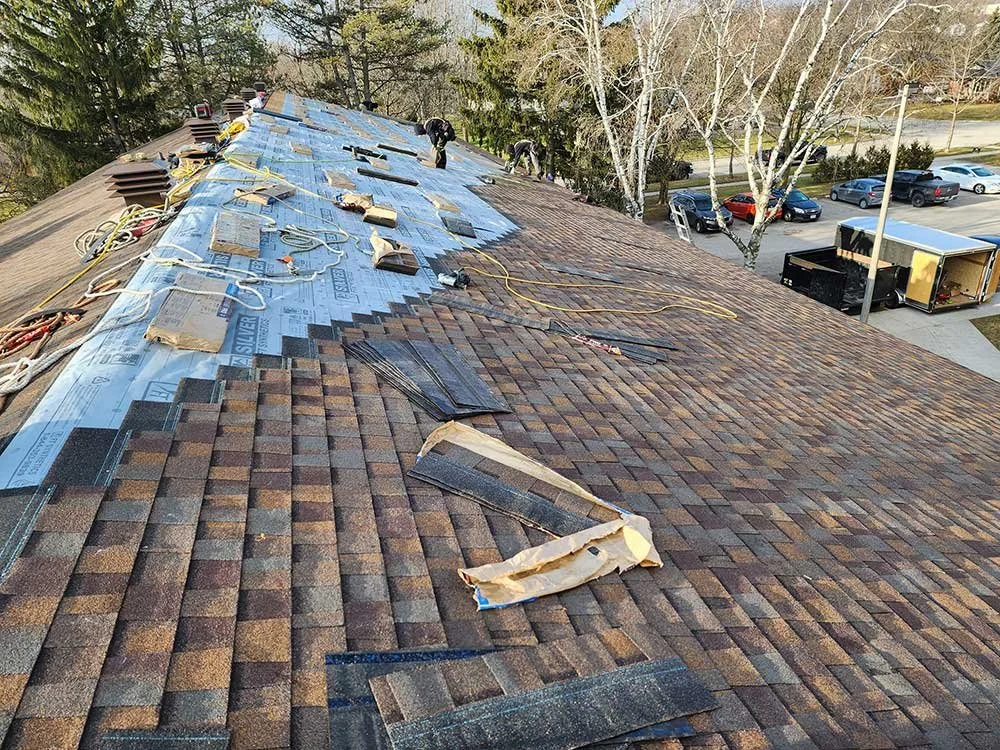 Roofing workers installing new shingles on a rooftop with tools and materials laid out, trees and a parking lot with cars in the background.