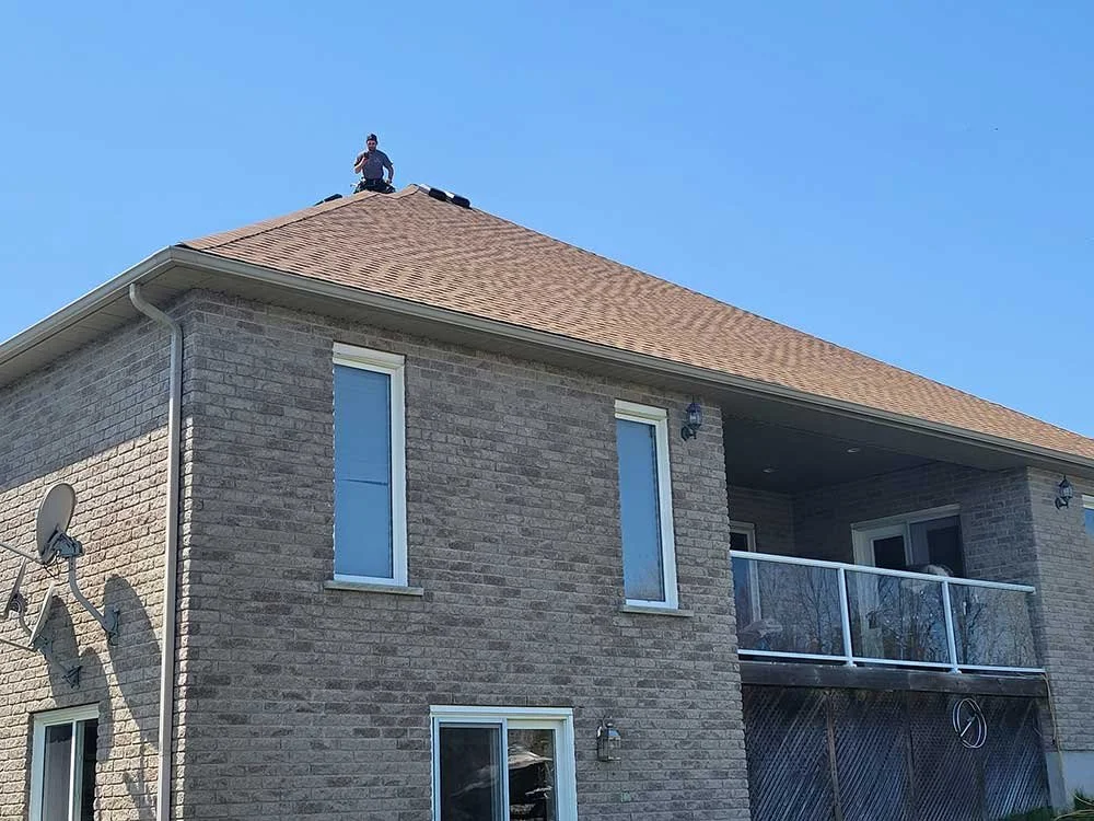 Person sitting on the roof of a two-story brick house on a sunny day. Quality Care Roofing is a Kitchener-Waterloo roofing company and roof repair contractor.