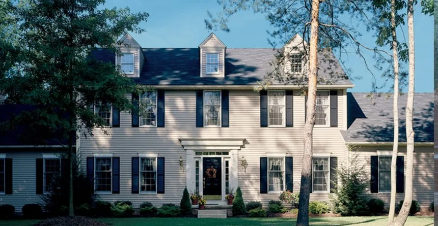 A large, two-story suburban house with white siding, black shutters, and dormer windows, surrounded by trees and a well-kept lawn.