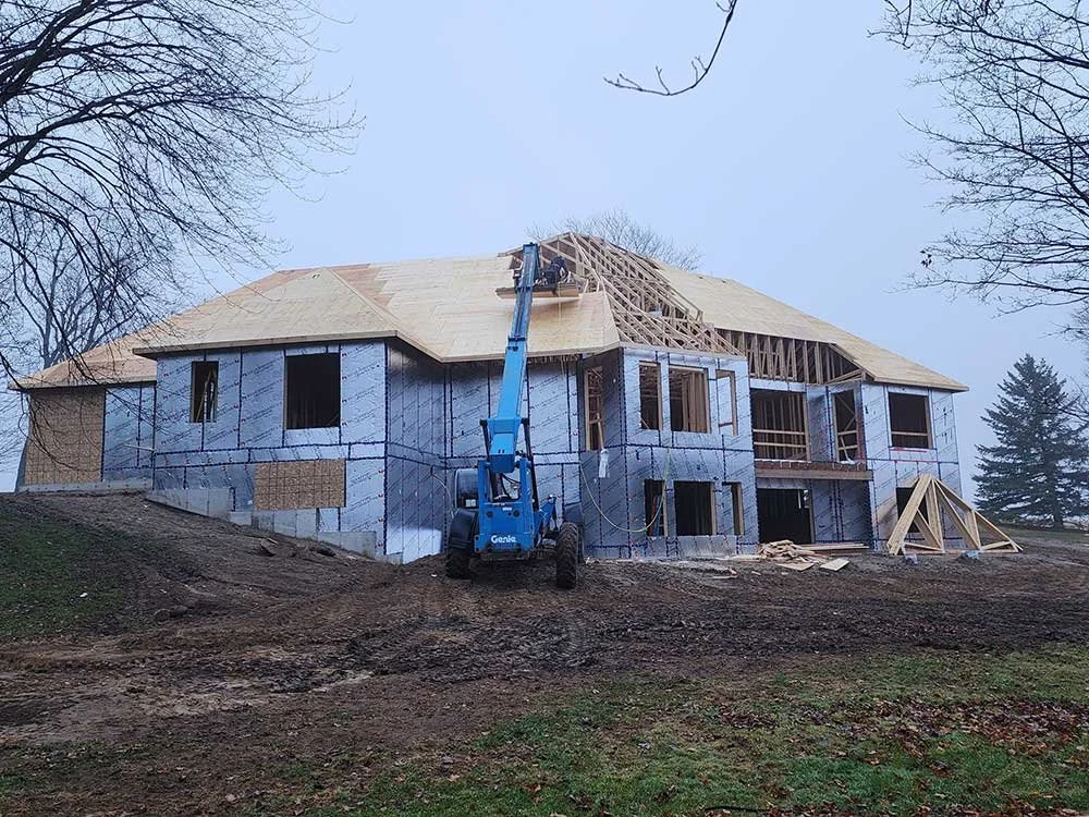 A house under construction with blue insulation and wooden framing, a blue lift platform is being used on the roof, and there are trees surrounding the site.