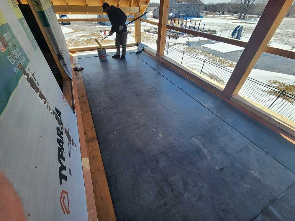A construction worker standing on a partially finished balcony floor, applying a black waterproofing membrane. The balcony is framed with wooden beams, with an outdoor scene including snow, trees, and playground equipment visible in the background.